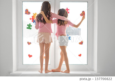 Two girls standing by a bright window decorating glass with colorful puzzle pieces and hearts. Autism awareness, friendship, childhood support, inclusion and neurodiversity concept. Two girls standing by a bright window decorating glass with colorful puzzle pieces and hearts. Autism awareness, friendship, childhood support, inclusion and neurodiversity concept. 137700822