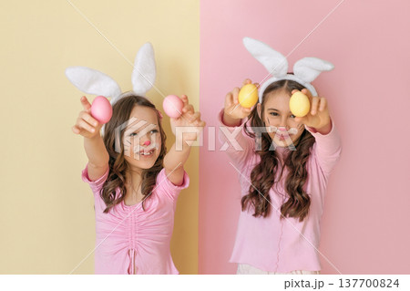 Two smiling girls wearing bunny ears and face paint holding colorful Easter eggs toward the camera on pastel background. Spring holiday, childhood joy, Easter celebration and festive fun concept. Two smiling girls wearing bunny ears and face paint holding colorful Easter eggs toward the camera on pastel background. Spring holiday, childhood joy, Easter celebration and festive fun concept. 137700824