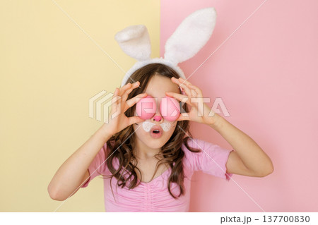 A surprised, whimsical girl hides her painted face behind Colored pink Easter eggs. A surprised, whimsical girl hides her painted face behind Colored pink Easter eggs. 137700830