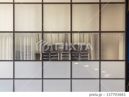 A study in quiet stillness, this tableau of functional simplicity is framed by a grid of glass and metal. Frosted lower panels obscure the floor, while clear panes above reveal utilitarian chairs 137703661