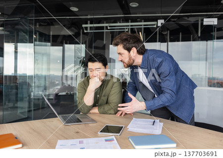 Two diverse male employees discussing a project, one appearing stressed and overwhelmed while the other expresses frustration and disagreement in a modern office meeting room setting Two diverse male employees discussing a project, one appearing stressed and overwhelmed while the other expresses frustration and disagreement in a modern office meeting room setting 137703691