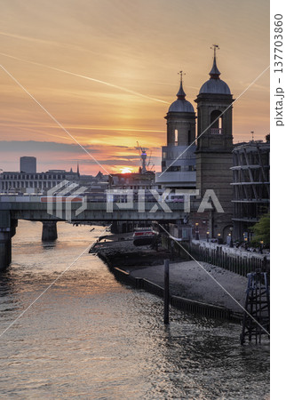 a breathtaking sunset over the River Thames in London, with the Cannon Street Railway Bridge spanning the water. The sky is ablaze with warm colors of orange, pink, and gold, which reflect beautifully 137703860