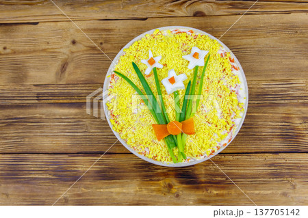 Traditional russian layered salad Mimosa with spring decoration Daffodil on wooden table. Top view. Decoration is made of egg, green onion and carrot Traditional russian layered salad Mimosa with spring decoration Daffodil on wooden table. Top view. Decoration is made of egg, green onion and carrot 137705142
