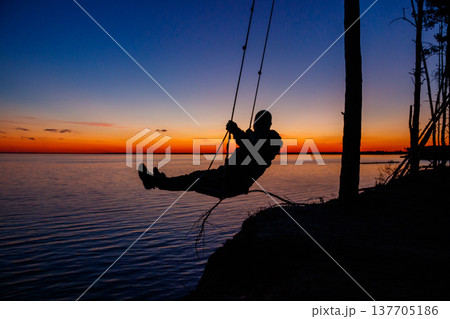Silhouette of a man on rope swing above river at sunset 137705186