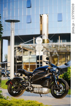 A vertical full-shot of a sleek black electric motorcycle parked on a sidewalk, showcasing its futuristic motor design against a backdrop of a modern glass building. A vertical full-shot of a sleek black electric motorcycle parked on a sidewalk, showcasing its futuristic motor design against a backdrop of a modern glass building. 137705839