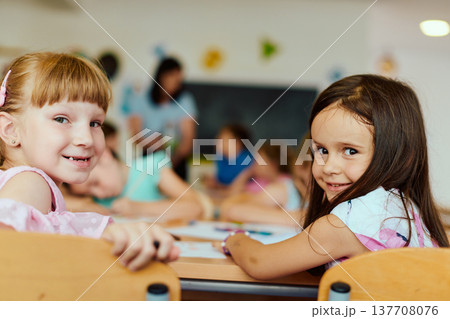 Two smiling girls sitting at a classroom table with classmates in the background 137708076