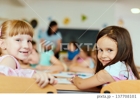 Two smiling girls in a bright classroom learning and drawing with classmates and a teacher nearby 137708101