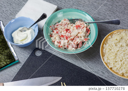 Various ingredients for sushi preparation are arranged on a kitchen countertop, featuring a bowl of crab salad, mayonnaise, rice, nori sheets, and utensils, creating a culinary setup Various ingredients for sushi preparation are arranged on a kitchen countertop, featuring a bowl of crab salad, mayonnaise, rice, nori sheets, and utensils, creating a culinary setup 137710278