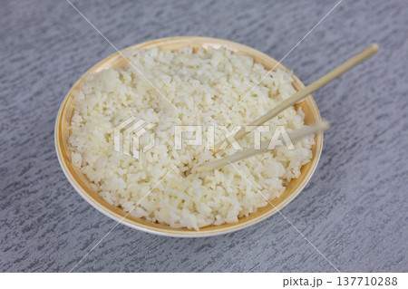Bowl filled with fluffy white rice, chopsticks positioned on top, set against a textured gray background, emphasizing the simplicity and cultural significance of rice in Asian cuisine Bowl filled with fluffy white rice, chopsticks positioned on top, set against a textured gray background, emphasizing the simplicity and cultural significance of rice in Asian cuisine 137710288