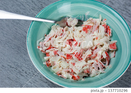 Bowl filled with crab salad featuring imitation crab meat, mayonnaise, and diced vegetables, placed on a textured gray surface with a fork positioned on the edge of the bowl 137710296