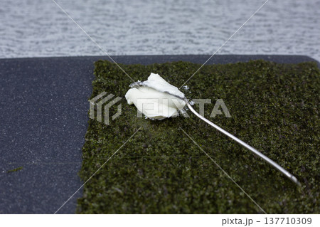 Close up of spoon with cream cheese on nori sheet. Essential step in sushi sandwich preparation, spreading creamy filling before adding rice and crab. Homemade Japanese cuisine process 137710309