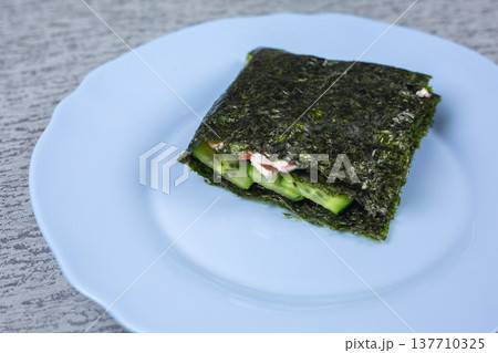 Seaweed wrap containing cucumber and cream cheese presented on a light blue plate, with a textured gray surface providing a contrasting backdrop for the dish Seaweed wrap containing cucumber and cream cheese presented on a light blue plate, with a textured gray surface providing a contrasting backdrop for the dish 137710325