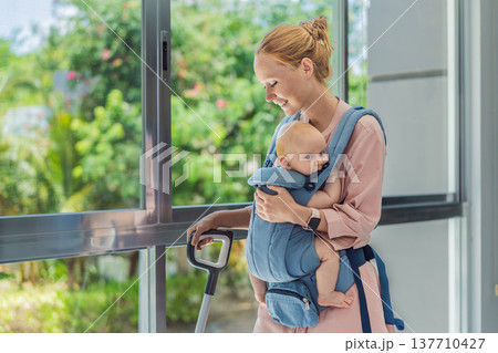 Woman vacuuming a room while carrying her baby in an ergonomic carrier. Modern motherhood, multitasking parent, home chores with infant, balance of childcare and housework concept. 137710427
