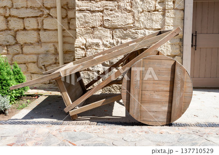 A wooden cart with a wheel is leaning against a stone wall 137710529