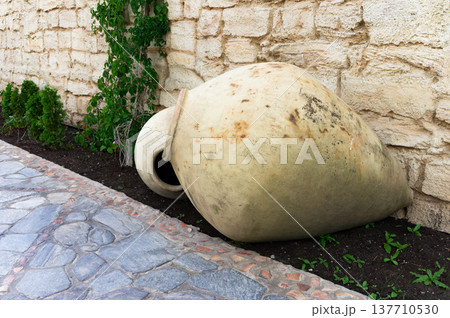 A large, brown, ceramic vase is sitting on the ground next to some plants 137710530