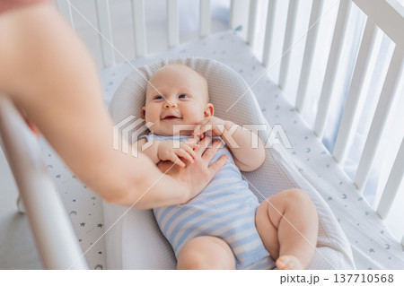 Mother gently sitting beside the crib and stroking her baby resting in a cozy cocoon nest. Tender intimate moment of care and trust at home. Motherhood, infant sleep and emotional bonding concept 137710568