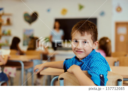 Smiling schoolboy sitting at desk in a bright classroom with teacher and classmates in background Smiling schoolboy sitting at desk in a bright classroom with teacher and classmates in background 137712085