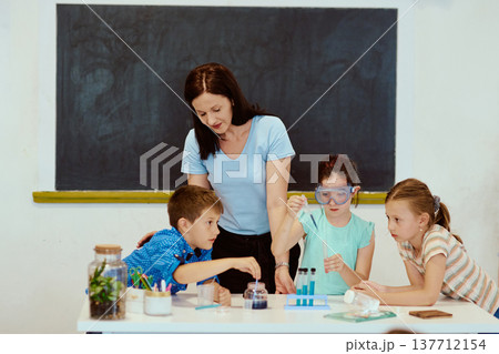 Teacher guides children through a hands-on science experiment in classroom with test tubes and goggles 137712154