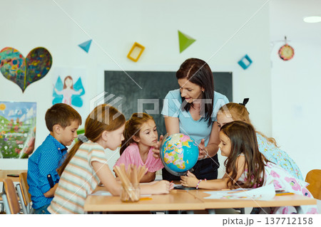 Teacher showing a globe to a group of curious children in a bright classroom learning together Teacher showing a globe to a group of curious children in a bright classroom learning together 137712158