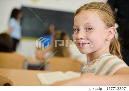 Smiling schoolgirl in classroom looking over shoulder during lesson with blurred teacher and classmates Smiling schoolgirl in classroom looking over shoulder during lesson with blurred teacher and classmates 137712207