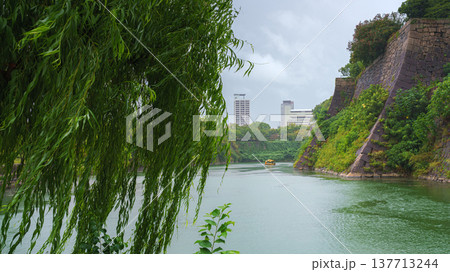 Osaka, Japan - Sep 22 2024, panoramic view of the moat around Osaka Castle on which a yellow tourist boat floats, view from the bridge through foliage and a tree branch, on a rainy day, Osaka, Japan 137713244