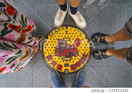 Osaka, Japan - Sep 22 2024, An overhead view of a manhole cover withred and yellow painting of fire truck, surrounded by the feet of four people, a man, a woman and a child, Osaka, Japan 137713291