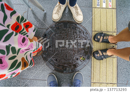 Osaka, Japan - Sep 22 2024, close-up view of metal manhole with patterns depicting a bridge over a river, buildings and a pagoda, surrounded by legs of four people, at Dotonbori district, Osaka, Japan Osaka, Japan - Sep 22 2024, close-up view of metal manhole with patterns depicting a bridge over a river, buildings and a pagoda, surrounded by legs of four people, at Dotonbori district, Osaka, Japan 137713293