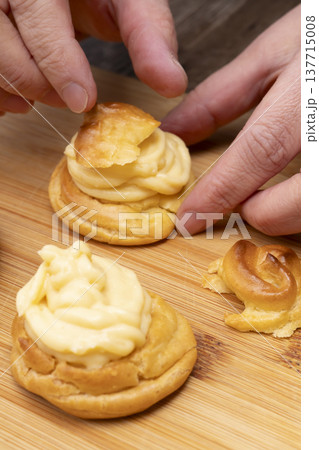 キッチンでシュークリームを作る手元 シュー生地にカスタードクリームを絞り粉糖をふるう キッチンでシュークリームを作る手元 シュー生地にカスタードクリームを絞り粉糖をふるう 137715008