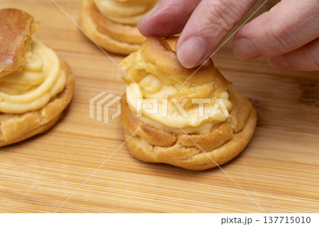 キッチンでシュークリームを作る手元　シュー生地にカスタードクリームを絞り粉糖をふるう 137715010