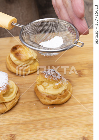 キッチンでシュークリームを作る手元　シュー生地にカスタードクリームを絞り粉糖をふるう 137715013