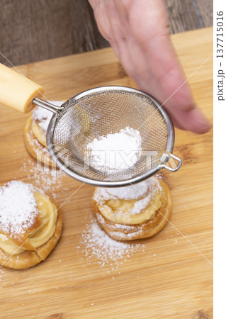 キッチンでシュークリームを作る手元　シュー生地にカスタードクリームを絞り粉糖をふるう 137715016
