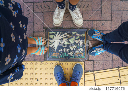 Himeji, Japan - Sep 24 2024, A close-up view of a manhole with colorful picture of stylized flying white egrets over a river and a field of reeds, surrounded by feet of a family of four, Himeji, Japan 137716076