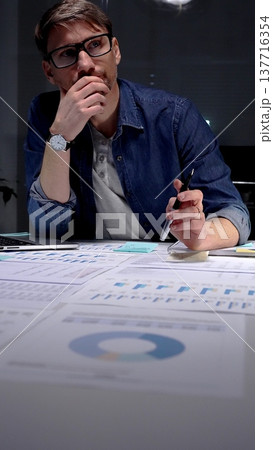 Professional man in glasses thinking deeply while analyzing financial charts and data on an office desk, working late night in a dark setting, feeling focused and determined 137716354