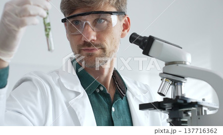 Adult male scientist in mask and gloves holding test tube with green plant sample, conducting laboratory research with a microscope. Medicine, healthcare and science concept Adult male scientist in mask and gloves holding test tube with green plant sample, conducting laboratory research with a microscope. Medicine, healthcare and science concept 137716362