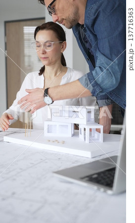Architects collaborating on building design, a female showing a colleague a blueprint layout while creating a physical architectural model in an office Architects collaborating on building design, a female showing a colleague a blueprint layout while creating a physical architectural model in an office 137716363