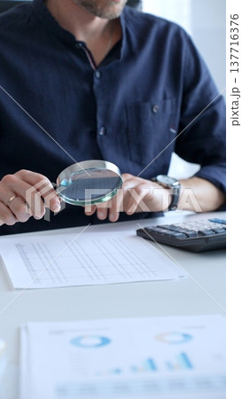 A financial analyst reviewing paperwork, using calculator and magnifying glass while examining corporate budget reports in modern workspace, close up. Audit and taxes, business concept A financial analyst reviewing paperwork, using calculator and magnifying glass while examining corporate budget reports in modern workspace, close up. Audit and taxes, business concept 137716376
