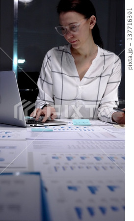 Businesswoman working late night in a modern office, analyzing financial data on a digital tablet and reviewing paperwork, demonstrating dedication and overtime productivity Businesswoman working late night in a modern office, analyzing financial data on a digital tablet and reviewing paperwork, demonstrating dedication and overtime productivity 137716391