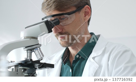 Senior man scientist wearing protective glasses and lab coat analyzing samples with a microscope in laboratory setting. Medicine, health care and science concept 137716392