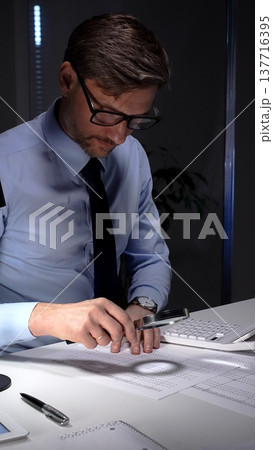 Businessman diligently auditing financial documents with a magnifying glass under focused lamp light. Audit and taxes in business Businessman diligently auditing financial documents with a magnifying glass under focused lamp light. Audit and taxes in business 137716395