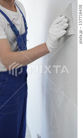 Close-up of man construction worker wearing gloves and blue overalls is using a putty knife to apply plaster on a white wall, performing home renovation and improvement Close-up of man construction worker wearing gloves and blue overalls is using a putty knife to apply plaster on a white wall, performing home renovation and improvement 137716438