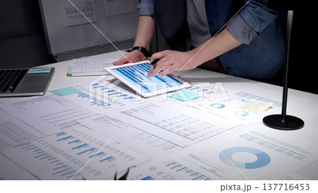 Person working late, analyzing financial data, charts, and graphs spread across a desk, using a laptop to review business analytics and market performance statistics Person working late, analyzing financial data, charts, and graphs spread across a desk, using a laptop to review business analytics and market performance statistics 137716453