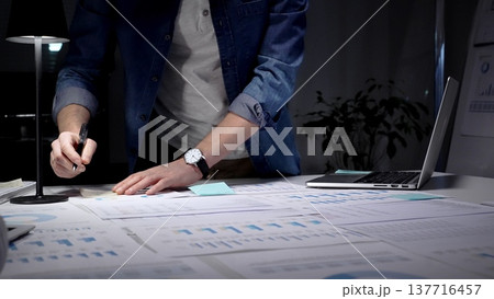 Entrepreneur standing at a desk in a dark office, studying financial reports and charts by lamp light while working on a laptop, focused on strategic planning and data analysis Entrepreneur standing at a desk in a dark office, studying financial reports and charts by lamp light while working on a laptop, focused on strategic planning and data analysis 137716457