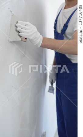 Unknown man construction worker wearing gloves and blue overalls is using a putty knife to apply plaster on a white wall, performing home renovation and improvement Unknown man construction worker wearing gloves and blue overalls is using a putty knife to apply plaster on a white wall, performing home renovation and improvement 137716488