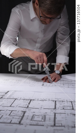 Architect working on blueprints using a compass, designing building plans in a studio at night. Concept of architecture and business in low key lighting Architect working on blueprints using a compass, designing building plans in a studio at night. Concept of architecture and business in low key lighting 137716504