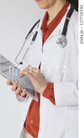 Unknown professional woman doctor, wearing white coat over a brick-red blouse and a stethoscope, is using a digital tablet, suggesting innovative healthcare practices in medicine. Health care concept Unknown professional woman doctor, wearing white coat over a brick-red blouse and a stethoscope, is using a digital tablet, suggesting innovative healthcare practices in medicine. Health care concept 137716592
