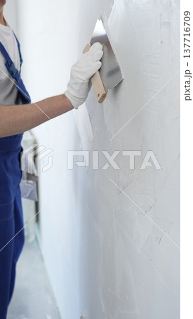 Close up of the construction worker hand wearing protective gloves, spreading plaster smoothly across wall using professional drywall taping knife realizing renovation project, vertical view Close up of the construction worker hand wearing protective gloves, spreading plaster smoothly across wall using professional drywall taping knife realizing renovation project, vertical view 137716709