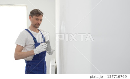 Middle aged male construction worker wearing gloves and blue overalls is using a putty knife to apply plaster on a white wall, performing home renovation Middle aged male construction worker wearing gloves and blue overalls is using a putty knife to apply plaster on a white wall, performing home renovation 137716769