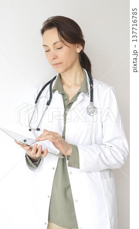 Portrait of a doctor woman wearing white medical coat over green blouse, stethoscope, is reviewing patient records on digital tablet, standing in medical workspace with natural lighting. Medicine Portrait of a doctor woman wearing white medical coat over green blouse, stethoscope, is reviewing patient records on digital tablet, standing in medical workspace with natural lighting. Medicine 137716785