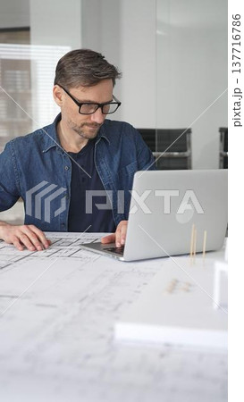 Male architect in glasses at a contemporary office working on a white architectural scale model of a modern house design, developing creative housing plans on a desk with blueprints and a laptop 137716786