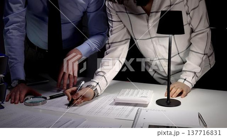 Business professionals analyzing financial data with a magnifying glass and calculator, focusing on document details and reviewing report figures during a late night work session 137716831